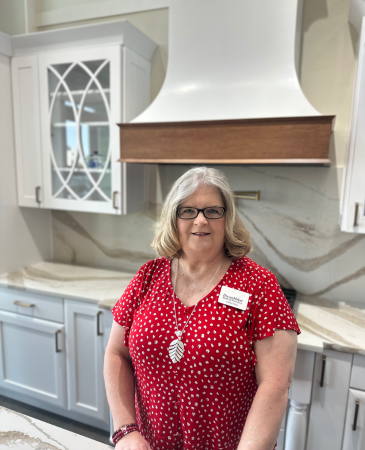 A smiling woman with shoulder-length blonde hair and glasses stands in a modern kitchen showroom, wearing a red patterned blouse and a name tag that reads “Debbie – DreamMaker Bath & Kitchen.”
